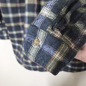 May include: Close-up of a blue, green, and tan plaid flannel shirt sleeve. The sleeve is buttoned at the cuff with a small, round, tan button. The fabric has a soft, textured appearance.