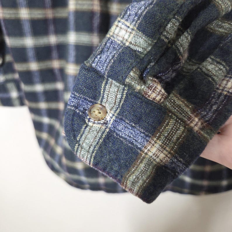 May include: Close-up of a blue, green, and tan plaid flannel shirt sleeve. The sleeve is buttoned at the cuff with a small, round, tan button. The fabric has a soft, textured appearance.