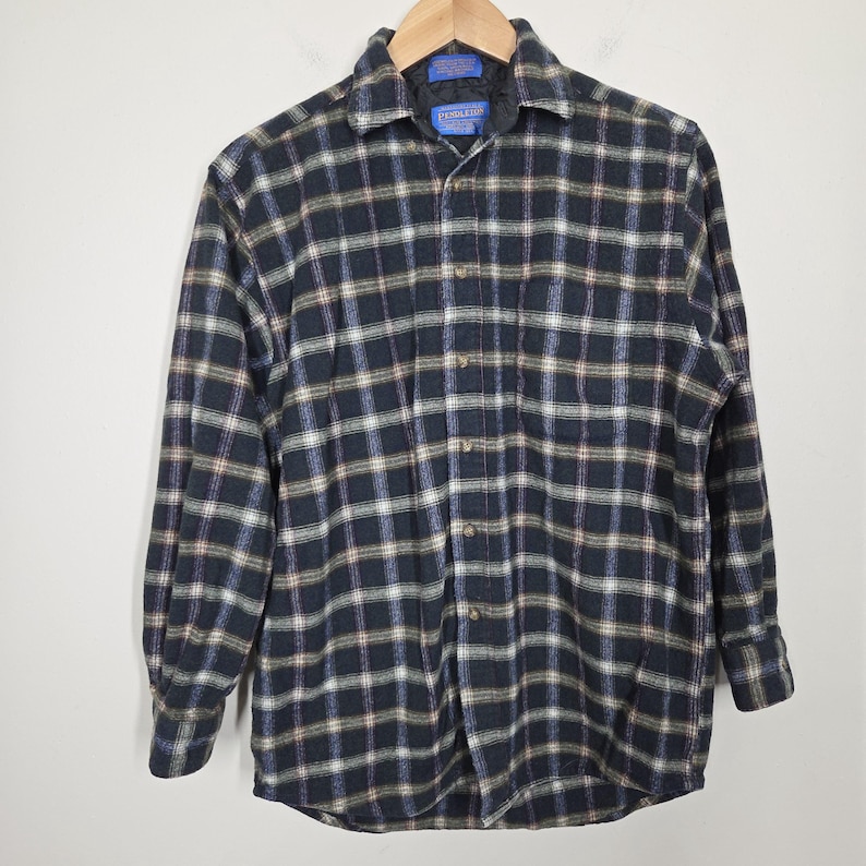 May include: A long-sleeved, button-down flannel shirt in a plaid pattern of navy blue, white, brown, and light blue. The shirt is hanging on a wooden hanger against a white background. The shirt has a collar and buttoned cuffs.