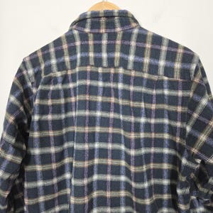 May include: A dark blue plaid flannel shirt with a button-down collar, hanging on a wooden hanger. The plaid pattern includes shades of blue, brown, and white. The shirt is made of a soft, woven fabric.