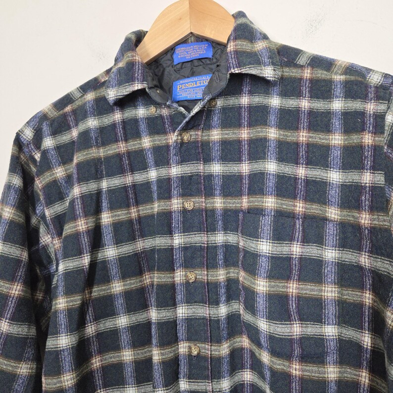 May include: A long-sleeved plaid shirt in shades of navy, green, white, and purple. The shirt has a button-down front, a collar, and a chest pocket. The brand name "PENDLETON" is visible on a tag near the collar.
