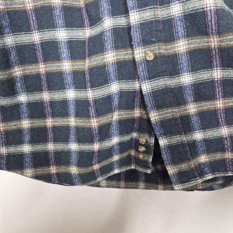 May include: Close-up of a button-down shirt with a plaid pattern in shades of navy, blue, white, and brown. The shirt has a curved hem and visible buttons. The fabric appears to be a soft, woven material.