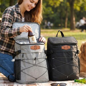 May include: A woman opening a gray and black insulated backpack cooler with a brown leather patch. A black insulated backpack cooler is next to it. A golden retriever dog is sitting nearby. Beer bottles and sunglasses are on the ground.