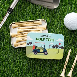 Op de afbeelding: Een golfcadeauset met een blikje houten golftees en een golfbal. Het blikje is versierd met een golfkar-illustratie en de tekst "RIVER'S GOLF TEES". De tees zijn gegraveerd met "RIVER'S TEES". De set ligt op groen gras.
