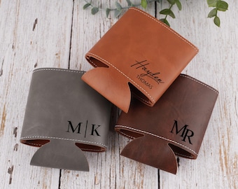 May include: Three personalized leather drink holders in shades of brown and gray are displayed. The drink holders have white stitching and are customized with initials or names. The background is a light wood surface.