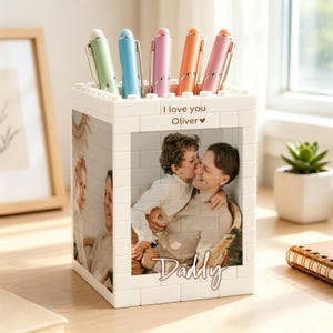 Puede incluir: Portalápices blanco estilo ladrillo con fotos y los textos "I love you Oliver" y "Daddy". Contiene cinco bolígrafos en colores pastel: verde, azul, rosa, naranja y rosa. El portalápices está sobre una superficie de madera.