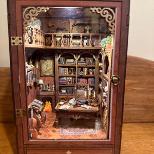 May include: A miniature wooden library diorama with intricate details. The interior features bookshelves, a desk, a fireplace, and a violin. The diorama is housed in a dark brown cabinet with ornate trim and a glass front, creating a three-dimensional scene.