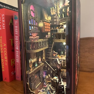 Könnte beinhalten: Ein detailliertes Miniatur-Diorama einer magischen Bibliothek oder eines Ladens mit mehreren Ebenen, Treppen und winzigen Figuren. Der Innenraum wird von kleinen Lichtern beleuchtet, die ein warmes Leuchten erzeugen. Die Außenseite ist eine dunkle, verzierte Box.