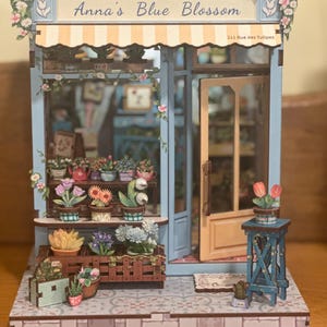 May include: A miniature model of a flower shop, "Anna's Blue Blossom," with a blue facade, striped awning, and open door. The shop is decorated with various potted flowers and greenery, with a small table holding tulips.
