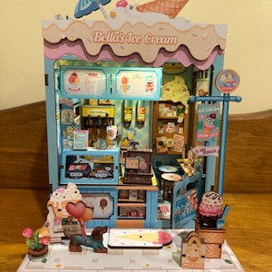 Könnte beinhalten: Miniatur-Eisdiele mit der Aufschrift "Bella's Ice Cream". Das Modell hat eine blau-weiße Farbgebung und zeigt detaillierte Innen- und Außenelemente, darunter Eistüten und eine Dackel-Figur.