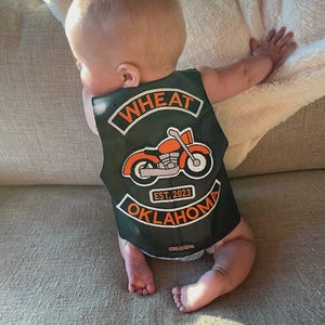 May include: A dark green baby vest featuring the words "WHEAT OKLAHOMA" and "EST. 2023" in white and orange. The vest has an orange motorcycle graphic.