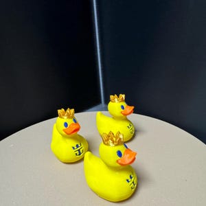 May include: Three yellow rubber duck figurines with gold crowns and orange beaks. Each duck has a blue logo on its side. The ducks are arranged on a light-colored surface against a dark background.