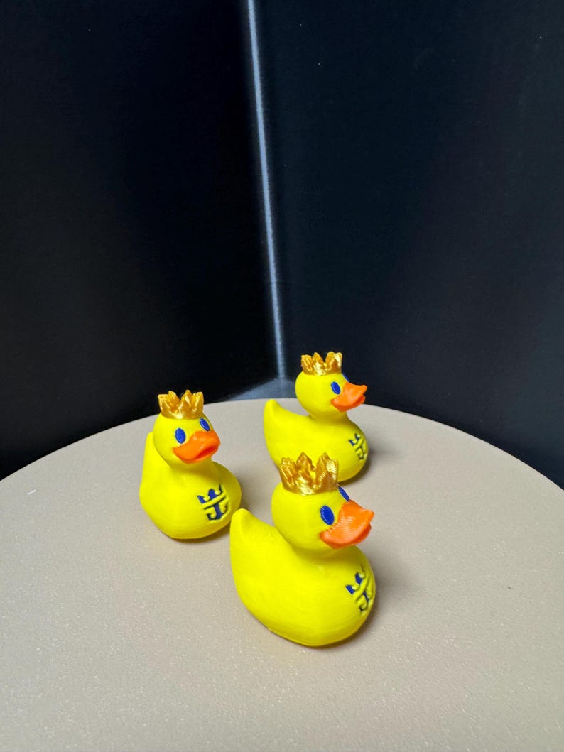 May include: Three yellow rubber duck figurines with gold crowns and orange beaks. Each duck has a blue logo on its side. The ducks are arranged on a light-colored surface against a dark background.