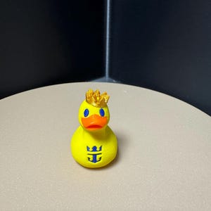 May include: A yellow rubber duck with a gold crown, blue eyes, and an orange beak. The duck has a blue logo on its chest. The duck is a novelty item.