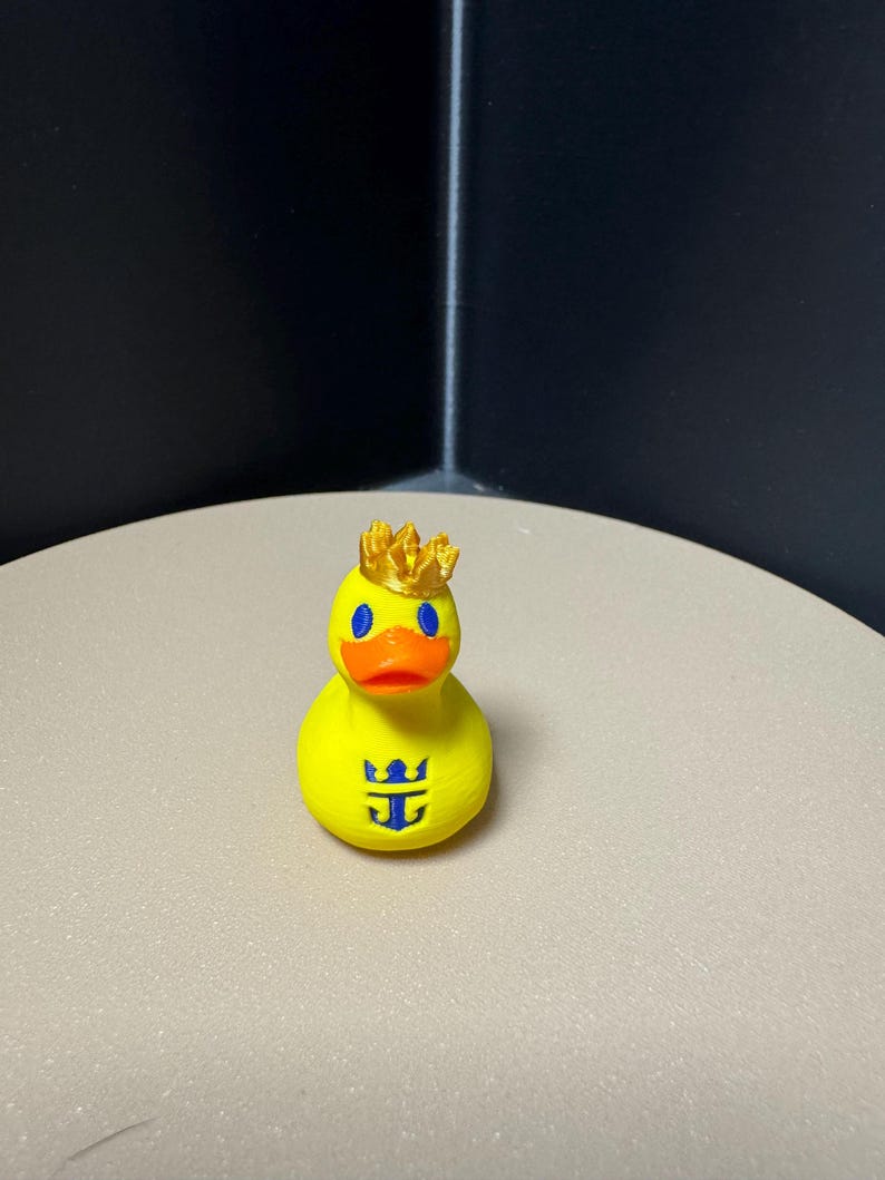 May include: A yellow rubber duck with a gold crown, blue eyes, and an orange beak. The duck has a blue logo on its chest. The duck is a novelty item.