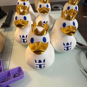 May include: A collection of white duck figurines, each adorned with a gold crown and a blue logo. The ducks have gold beaks and blue eyes. The figurines are arranged on a work surface.
