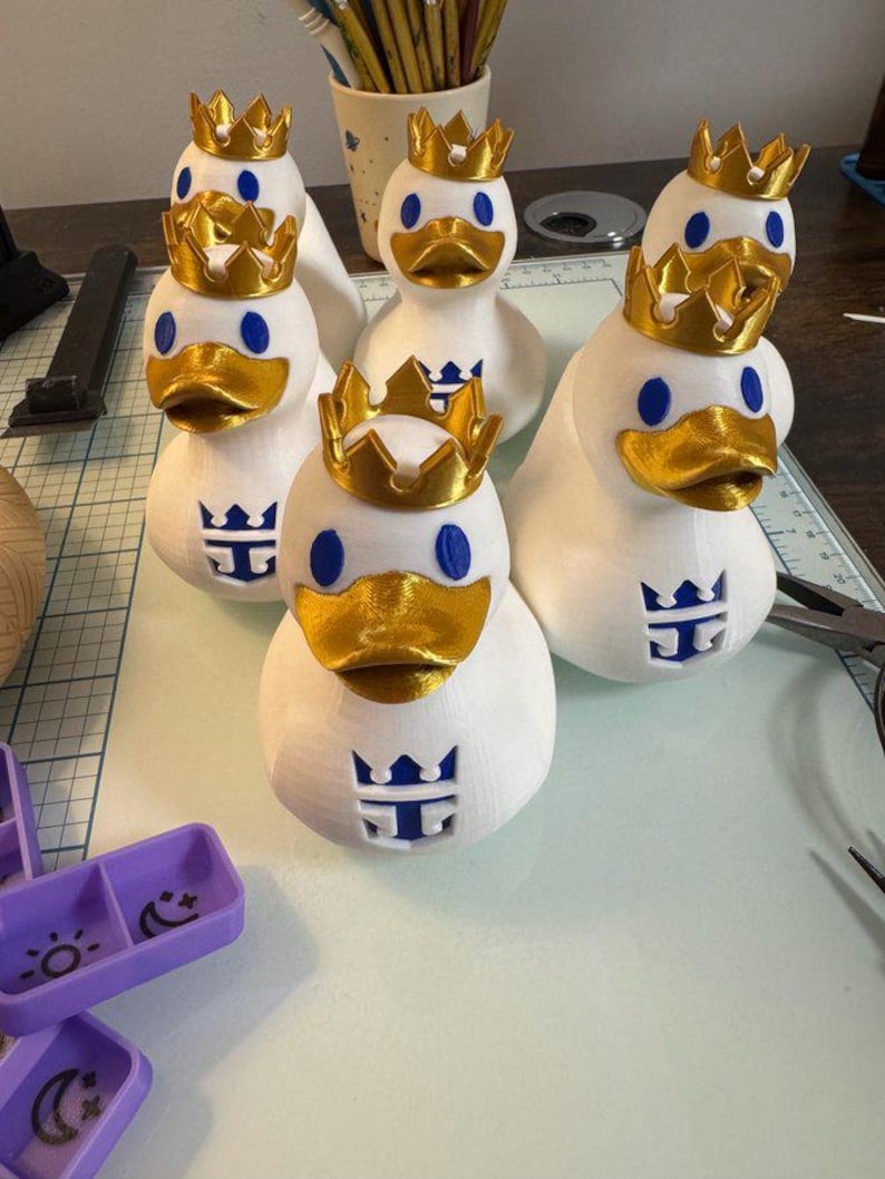 May include: A collection of white duck figurines, each adorned with a gold crown and a blue logo. The ducks have gold beaks and blue eyes. The figurines are arranged on a work surface.
