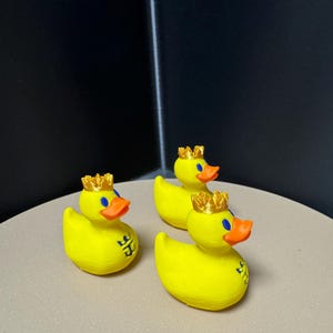 May include: Three yellow rubber duck figurines with orange beaks, blue eyes, and gold crowns. Each duck has a small blue emblem on its side. The ducks are arranged on a light brown surface against a dark background.