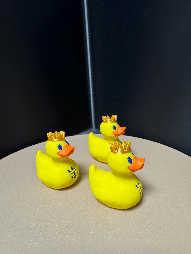 May include: Three yellow rubber duck figurines with orange beaks, blue eyes, and gold crowns. Each duck has a small blue emblem on its side. The ducks are arranged on a light brown surface against a dark background.