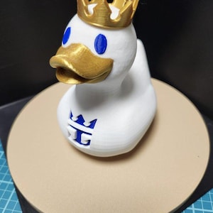 May include: A white duck figurine with a gold crown, beak, and blue eyes. The duck has a logo on its chest. The figurine is sitting on a tan circular base.