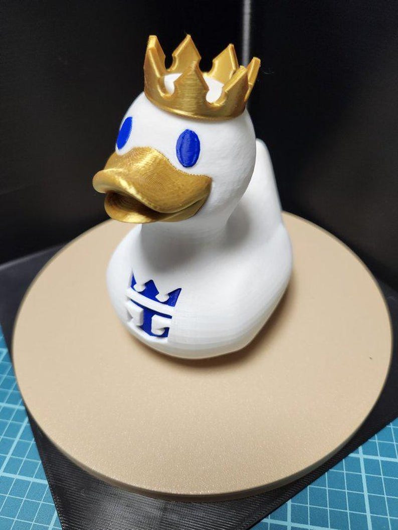 May include: A white duck figurine with a gold crown, beak, and blue eyes. The duck has a logo on its chest. The figurine is sitting on a tan circular base.