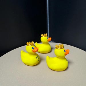 May include: Three yellow rubber duck figurines, each adorned with a gold crown and an orange beak. The ducks have blue eyes and a logo on their side. They are arranged on a light gray surface.