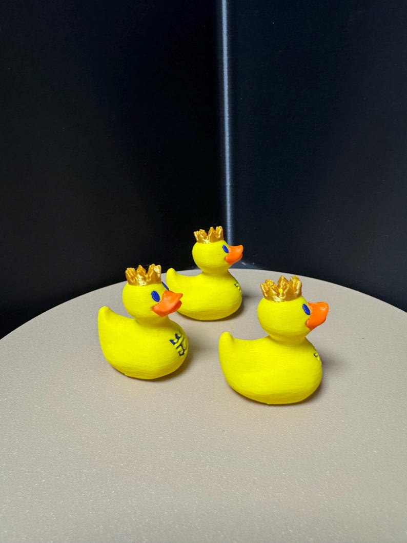 May include: Three yellow rubber duck figurines, each adorned with a gold crown and an orange beak. The ducks have blue eyes and a logo on their side. They are arranged on a light gray surface.