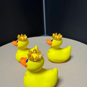 May include: Three yellow rubber duck figurines, each adorned with a gold crown. The ducks have orange beaks and blue eyes. The background is a neutral gray surface and a dark corner.