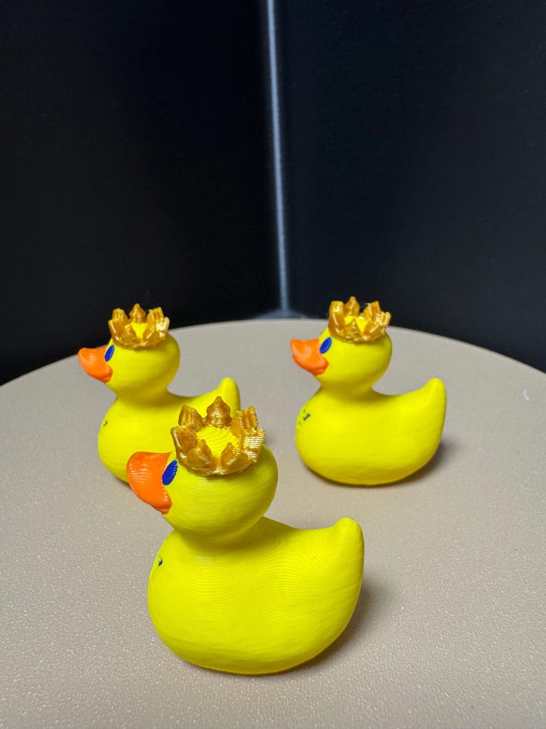 May include: Three yellow rubber duck figurines, each adorned with a gold crown. The ducks have orange beaks and blue eyes. The background is a neutral gray surface and a dark corner.