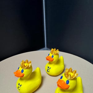 May include: Three yellow rubber duck figurines, each adorned with a gold crown and an orange beak. The ducks have blue eyes and a blue emblem on their chest. They are arranged on a light tan surface.