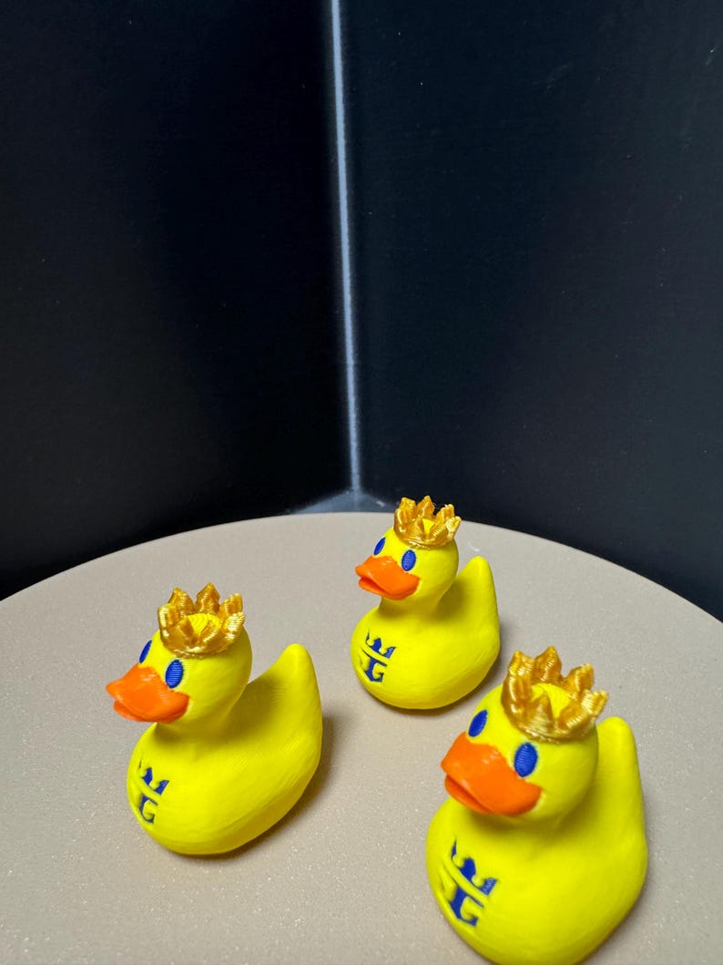 May include: Three yellow rubber duck figurines, each adorned with a gold crown and an orange beak. The ducks have blue eyes and a blue emblem on their chest. They are arranged on a light tan surface.