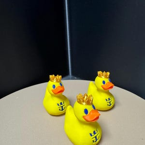May include: Three yellow rubber duck toys with gold crowns, blue eyes, and orange beaks. Each duck has a blue logo on its chest. The ducks are on a light tan surface with a dark background.