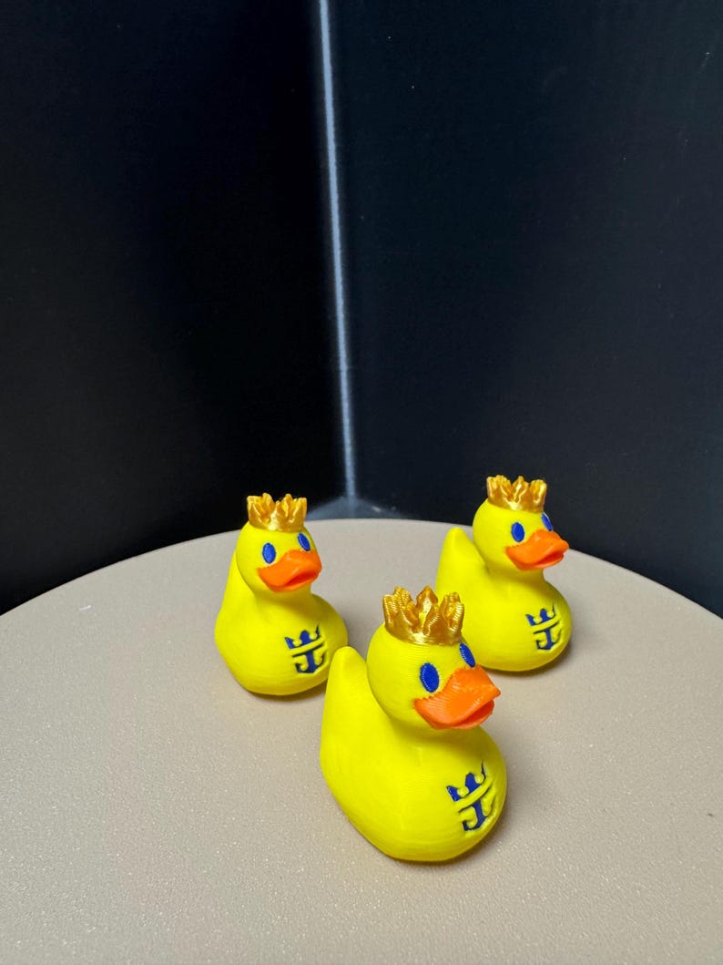 May include: Three yellow rubber duck toys with gold crowns, blue eyes, and orange beaks. Each duck has a blue logo on its chest. The ducks are on a light tan surface with a dark background.