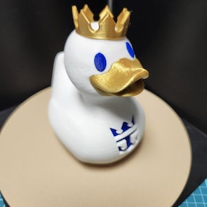 May include: A white duck figurine with a gold crown, beak, and blue eyes. The duck has a blue logo on its chest. The figurine is sitting on a beige circular surface.