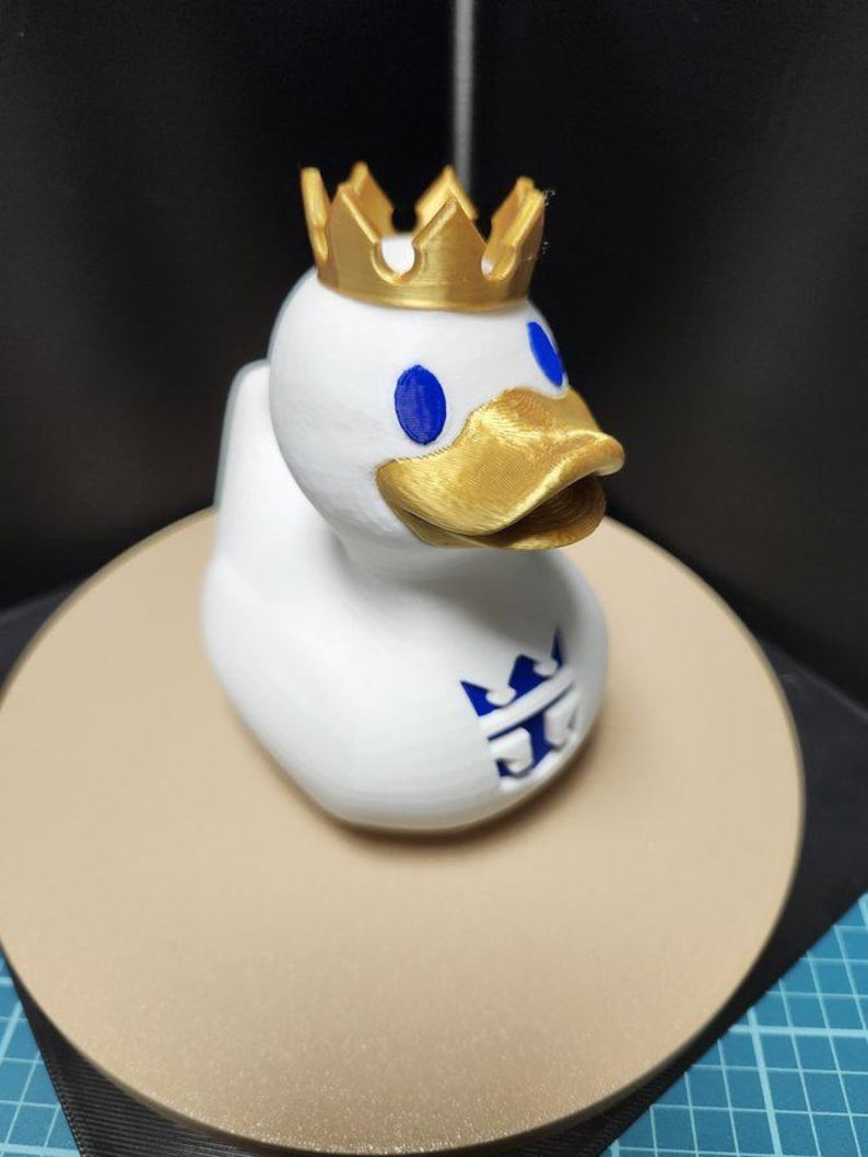 May include: A white duck figurine with a gold crown, beak, and blue eyes. The duck has a blue logo on its chest. The figurine is sitting on a beige circular surface.
