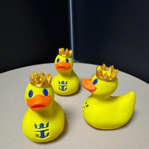 May include: Three yellow rubber duckies with orange beaks, blue eyes, and gold crowns. Each duck has a blue logo on its chest. The ducks are on a light-colored surface.