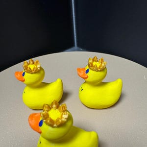 May include: Three yellow rubber duck figurines with orange beaks and blue eyes. Each duck wears a gold crown. The ducks are arranged on a light brown surface.