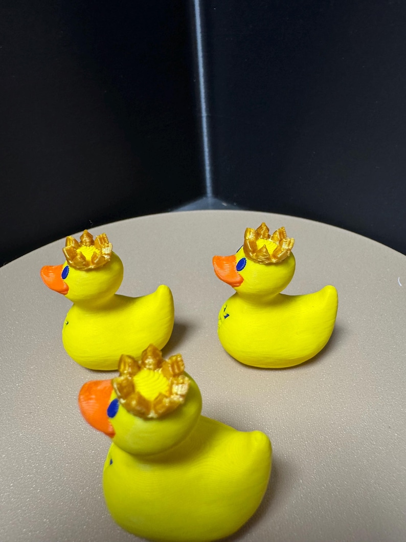 May include: Three yellow rubber duck figurines with orange beaks and blue eyes. Each duck wears a gold crown. The ducks are arranged on a light brown surface.