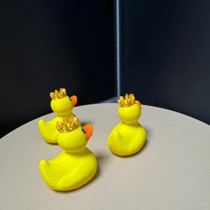 May include: Three yellow rubber duck figurines with orange beaks and gold crowns. The ducks are arranged on a light gray surface against a dark background. The ducks are a classic toy with a regal twist.