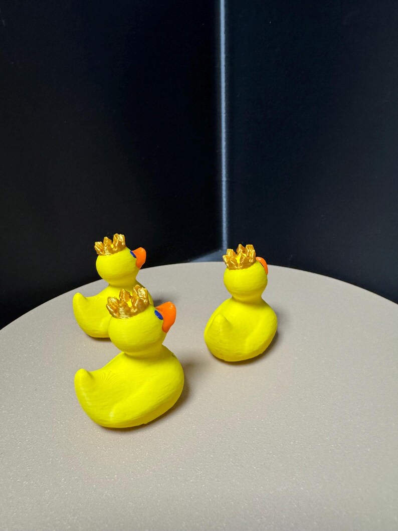 May include: Three yellow rubber duck figurines with orange beaks and gold crowns. The ducks are arranged on a light gray surface against a dark background. The ducks are a classic toy with a regal twist.