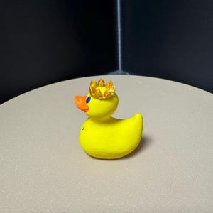 May include: A yellow rubber duck with an orange beak and a gold crown. The duck has blue eyes and is sitting on a light-colored surface. The duck is a novelty item.