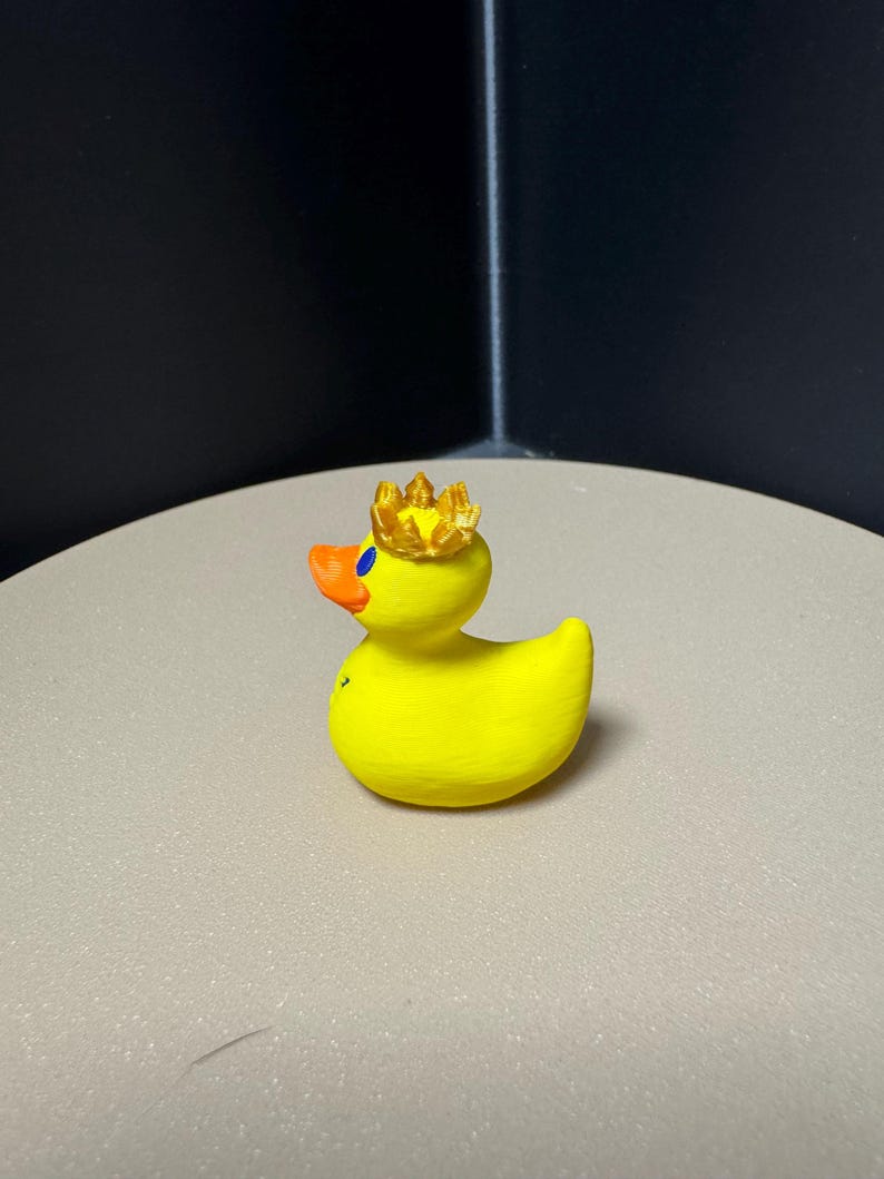 May include: A yellow rubber duck with an orange beak and a gold crown. The duck has blue eyes and is sitting on a light-colored surface. The duck is a novelty item.