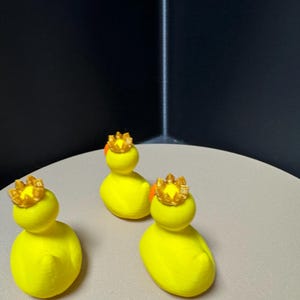 May include: Three yellow duck figurines, each wearing a gold crown. The ducks are positioned on a light-colored surface against a dark background. The crowns have an orange accent.