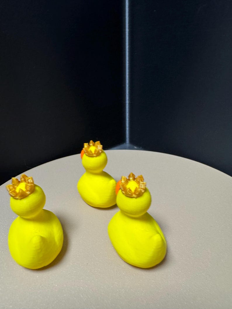 May include: Three yellow duck figurines, each wearing a gold crown. The ducks are positioned on a light-colored surface against a dark background. The crowns have an orange accent.