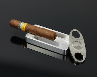 May include: A brown cigar with a yellow and black Cohiba band resting on a rectangular silver metal ashtray next to an oval-shaped silver metal cigar cutter with two finger holes and a central circular blade mechanism for cutting cigars.