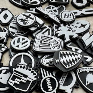 May include: A collection of black and white keyrings featuring various car logos, including Rover, Mini, Lamborghini, and VW. The keyrings are made of plastic and have a 3D design.