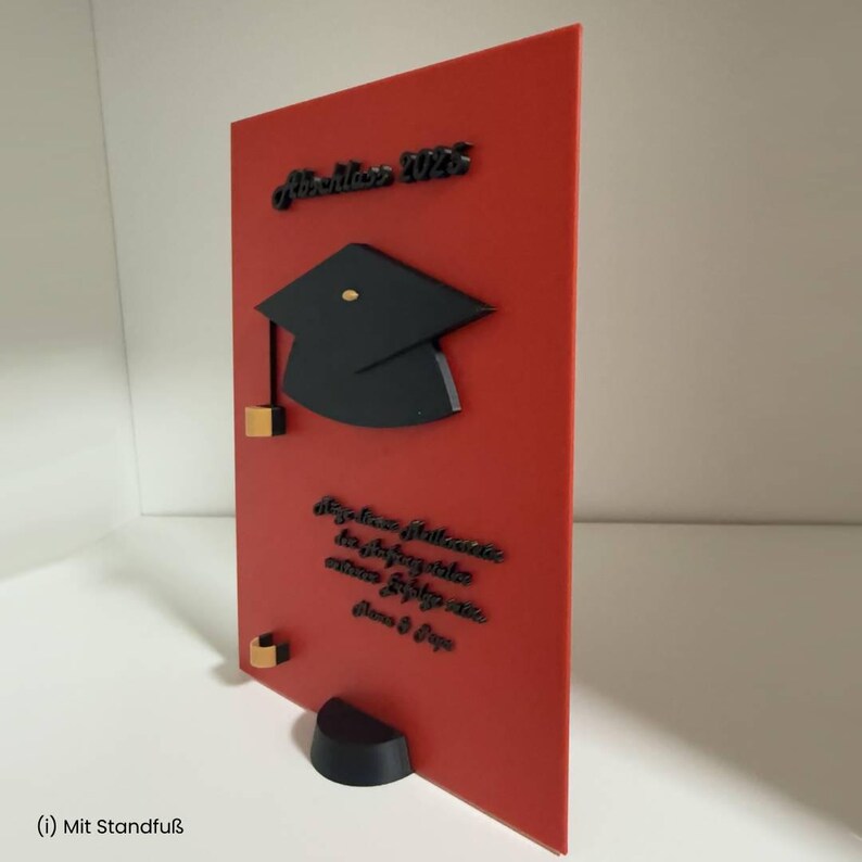 K&ouml;nnte beinhalten: Eine rechteckige rote Platte mit einem schwarzen Doktorhut-Symbol, dem Text "Abschluss 2028" in Schwarz und weiterem schwarzem Schrifttext darunter. Die Platte enth&auml;lt kleine goldfarbene Akzente und steht auf einem schwarzen, halbkreisf&ouml;rmigen Sockel.