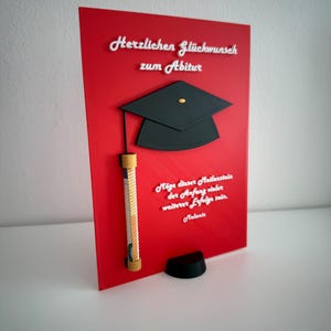 K&ouml;nnte beinhalten: Eine rechteckige rote Tafel mit einer schwarzen Doktorhut-Applikation, einem gerollten Geldschein in einer Halterung und wei&szlig;em erhabenem Text: Herzlichen Gl&uuml;ckwunsch zum Abitur, M&ouml;ge dieser Meilenstein der Anfang vieler weiterer Erfolge sein, Melanie.