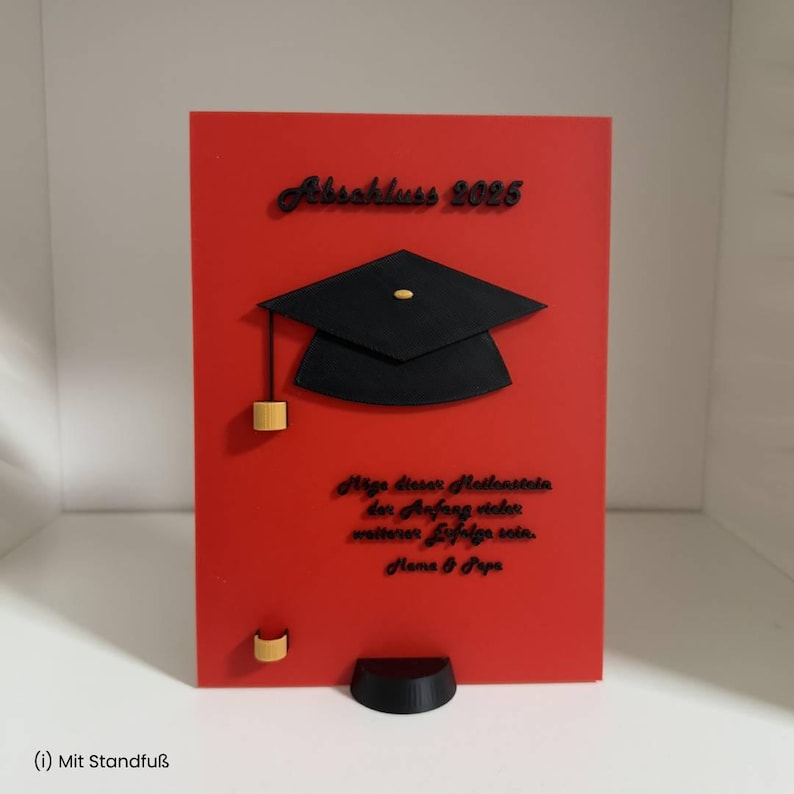 K&ouml;nnte beinhalten: Eine rechteckige rote Tafel mit einem schwarzen, erhabenen Doktorhut in der Mitte. Der Text lautet oben "Abschluss 2025", gefolgt von einer deutschen Inschrift und "Mama & Papa". An der Unterseite befindet sich ein schwarzer Standfu&szlig;.