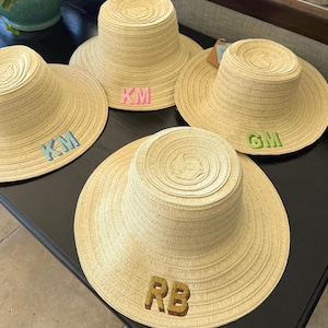 May include: Four light beige straw hats with flat tops and wide brims. Each hat features two-letter embroidered patches in different colors: light blue KM, pink KM, lime green GM, and gold RB. The hats are arranged in a group on a dark surface.